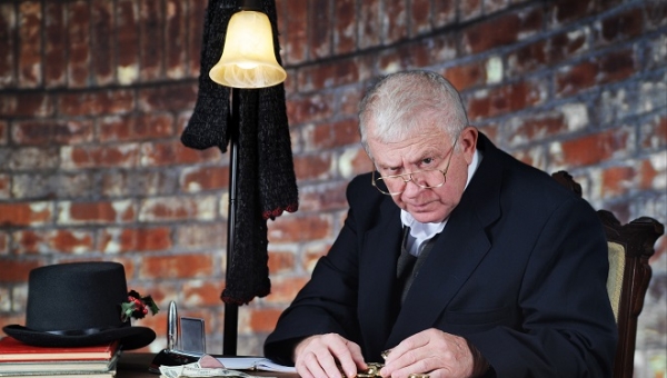 A grumpy old man scowling at the viewer as he counts his gold coins by a stack of large bills.